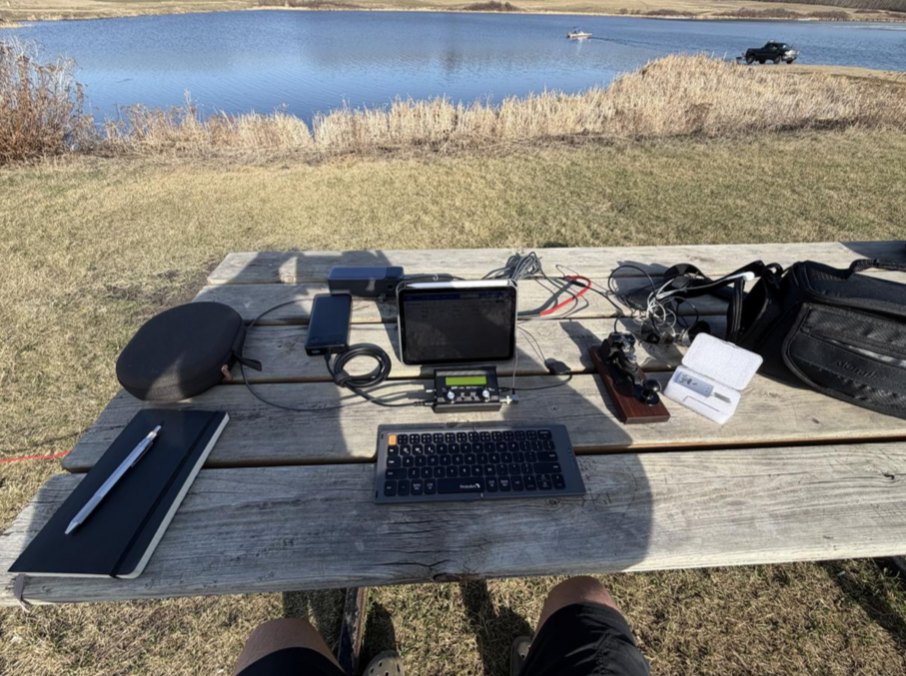 Portable CW station on a picnic table beside a lake: QMX, key, keyboard, tablet and logbook